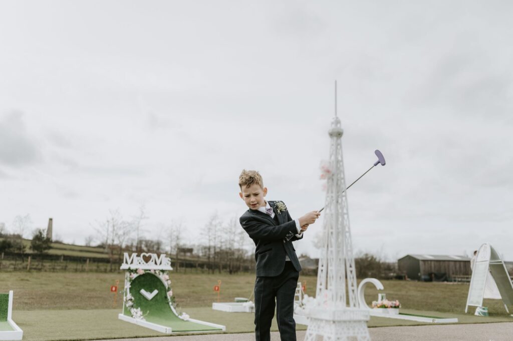 A young boy in a suit plays wedding mini golf outdoors, swinging a purple putter near a small Eiffel Tower replica. The course features decorative obstacles and a rural landscape in the background.