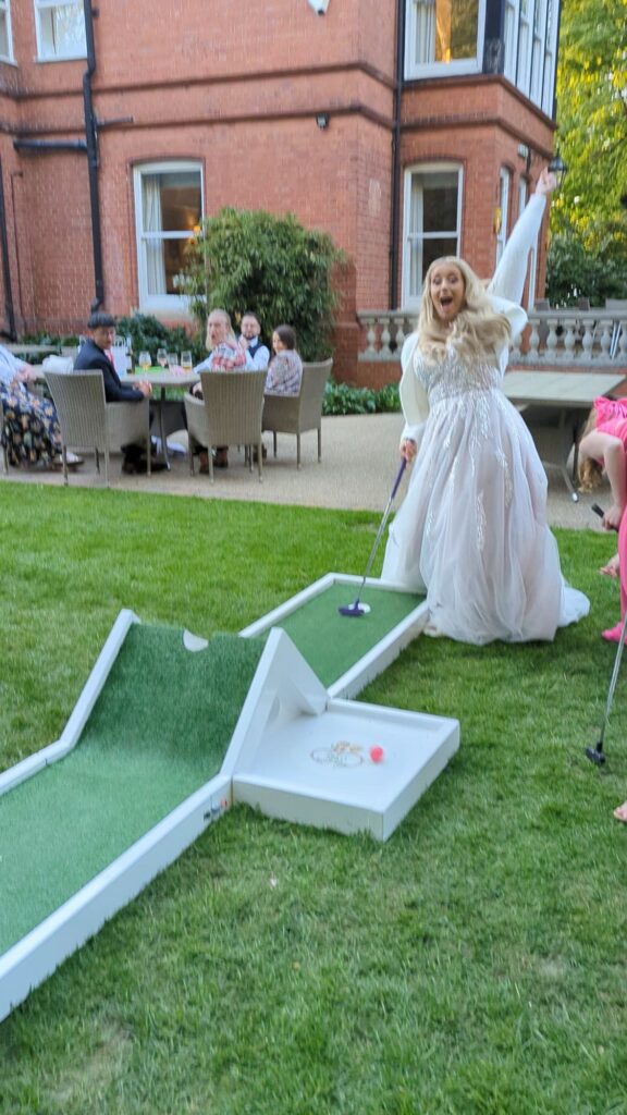 A bride in a white wedding dress excitedly plays wedding mini golf on a lawn, holding a putter near a small green course, while guests sit at tables outside a large brick building in the background.