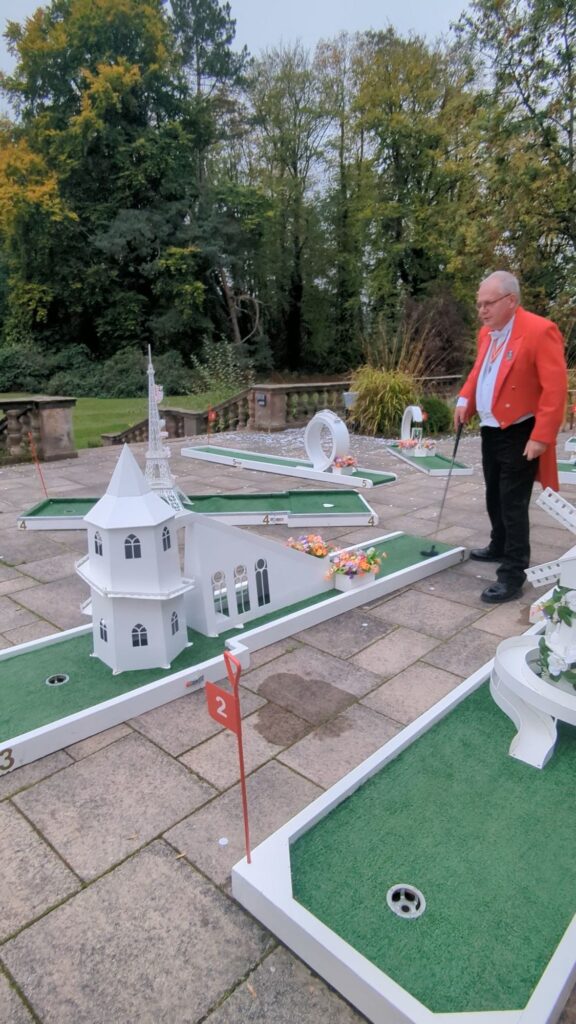 A toastmaster in a red jacket stands holding a mini-golf club on an outdoor wedding mini golf course including a castle, windmill, and Eiffel Tower. Lush trees and bushes are in the background.