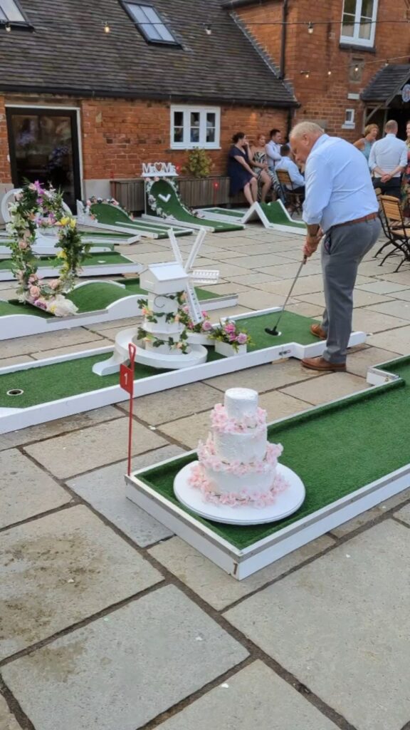 An older man enjoys a round of wedding mini golf on a course adorned with a giant tiered cake and floral arrangements, as a group of people looks on in the background.